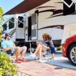 Mother father and son sitting near camping trailer smiling. Woman men kid relaxing on chairs near car and palms. Family spending time together on vacation near sea or ocean in modern rv park