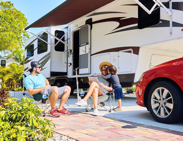 Mother father and son sitting near camping trailer smiling. Woman men kid relaxing on chairs near car and palms. Family spending time together on vacation near sea or ocean in modern rv park